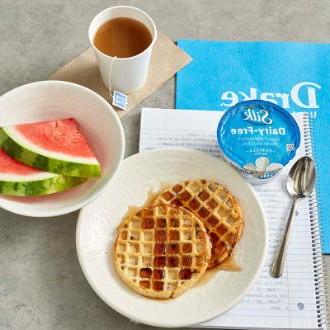 A cup of coffee next to two plates of waffles and watermelon sitting on top of a notebook on a table inside of a Drake University dining hall